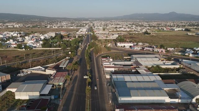 Puebla, mexico - 04.20.2026: Aerial flying forward along a multi lane highway running through a suburban town with industrial buildings and homes, revealing mountains on the horizon