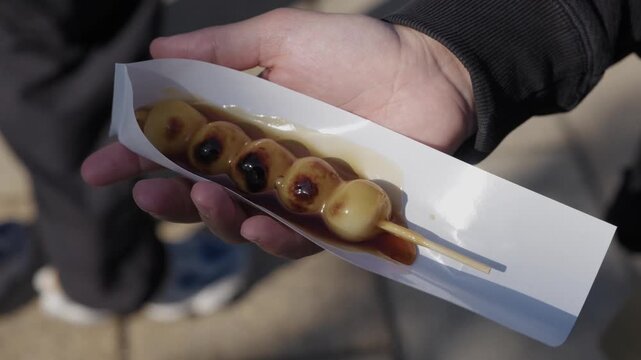 Hand holds skewer of grilled mitarashi dango rice dumplings with soy glaze in paper tray