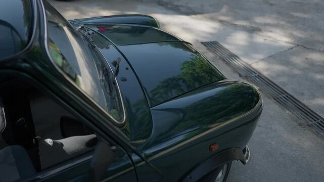 Polished dark green vintage car roof and rear glass reflecting trees on driveway