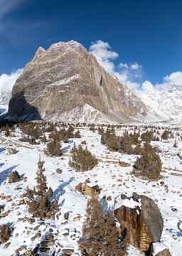 Aerial view of the snow-covered Hushe valley with a rocky mountain peak, sparse juniper trees, and deep shadows across the winter landscape in Khaplu, Gilgit Baltistan, Pakistan.