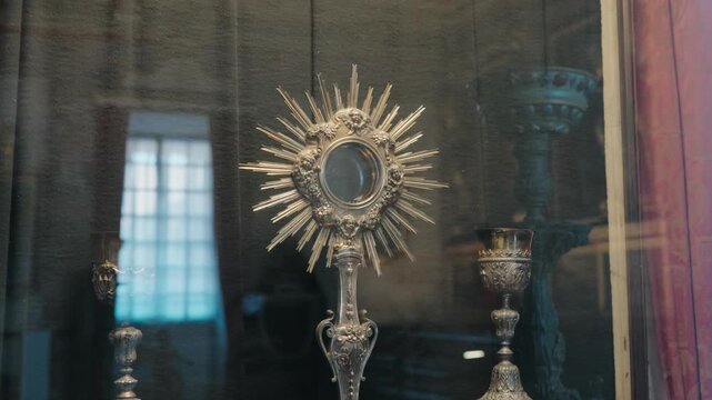 Gilded catholic ostensorium beside ornate silver chalice displayed behind glass