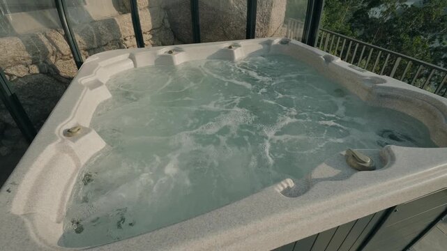Bubbling hot tub jacuzzi with swirling water on glass enclosed terrace beside stone wall