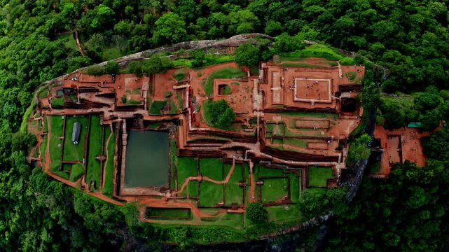 High aerial view of the Sigiriya rock fortress, showing the full plateau of ancient palace ruins, water cistern and gardens surrounded by tropical jungle.