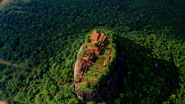 Aerial orbit of the ancient Sigiriya rock fortress summit, revealing weathered red-brick palace ruins and terraces perched high above dense green jungle.