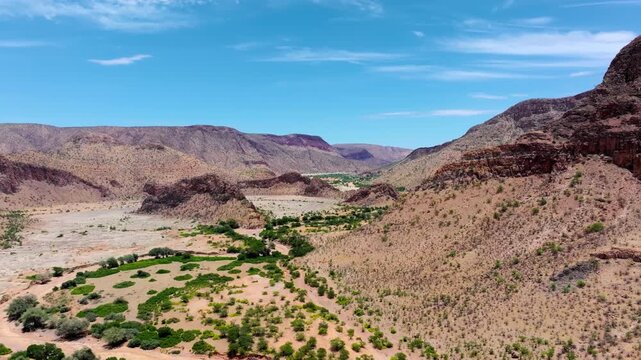 Low aerial flight along a green ephemeral riverbed winding between rugged ochre mountains in central Namibia, desert vegetation lining the banks.