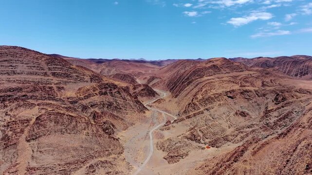 Drone flight above dramatically folded rock strata in the central Namibian desert, layered ridges stretching to a hazy horizon under a clear sky.