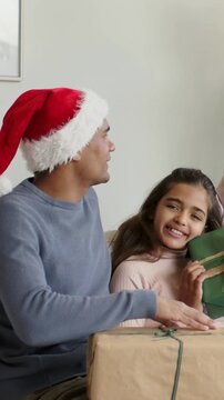 Vertical video: Noticing green gift, Santa-hatted father tickling girl on couch holding brown box