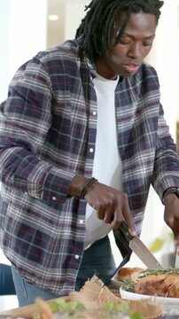 Vertical video: Leaning man in plaid carving roast at dining table with knife and fork serving guest