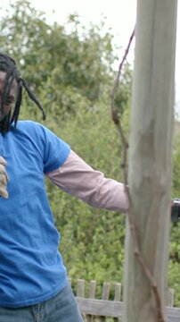 Vertical video: Reacting to gloved reach, man in blue tee laughing, reaching to post at garden, pot