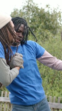 Vertical video: Clasping gloves two men in blue tees laughing and leaning by garden post after work