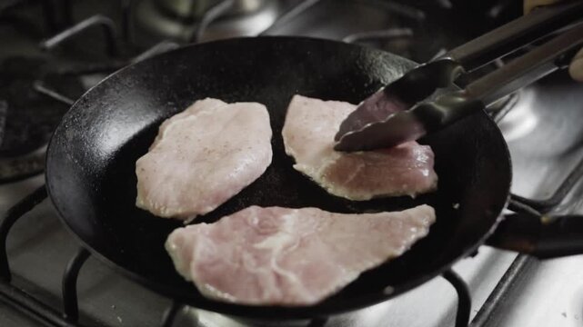 Close up shot of pork loin slices cooking in frying pan while being turned with kitchen tongs, showcasing meat browning process on gas stove during meal preparation