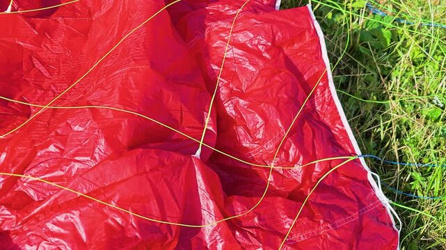 Detailed view of paragliding equipment on a grassy hillside, showing red parachute fabric, colorful control lines, stitching, and ripstop texture in bright summer sunlight before takeoff.