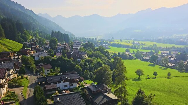 Elevated cable car view moving across Kitzbuhel, Austria, with village homes, rolling fields, dark forests, and layered blue mountains under strong afternoon sunlight and travel scenery.