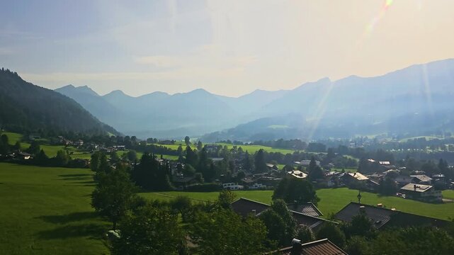 Wide aerial style view from a moving cable car, looking over village rooftops, rolling meadows, pine forests, and hazy mountain ridges under bright summer sun and lens flare.