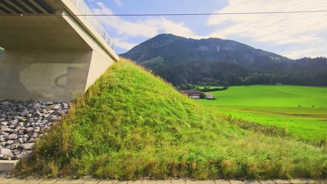 Train journey past open fields, scattered farm buildings, pine forests, and mountain hillsides, capturing peaceful countryside scenery from the window in peaceful rural scenery.