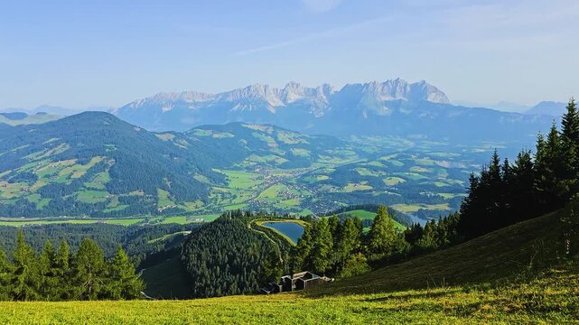Scenic view from above Kitzbuhel, Austria, showing bright green ski slopes, forested hills, valley towns, alpine lake, and dramatic Tyrolean Alps on a sunny summer travel day in the Alps.