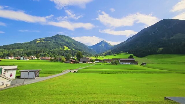 Austria rail travel through a peaceful mountain valley, showing bright meadows, chalet style homes, roadside trees, and forested peaks under summer skies.