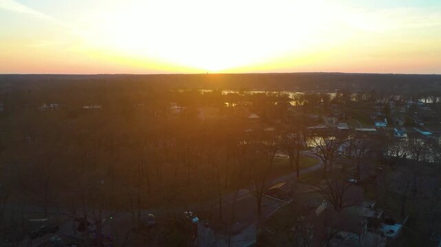 Wide aerial shot near Branson, Missouri, with bright sunset over calm Ozark lake water, forested shoreline, distant homes, soft haze, and golden reflections across the reservoir.