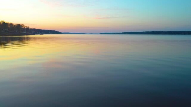 Scenic drone view near Branson, Missouri, gliding over calm lake water with pastel sunset sky, wooded Ozark hills, distant shoreline homes, and gentle reflections on the reservoir.