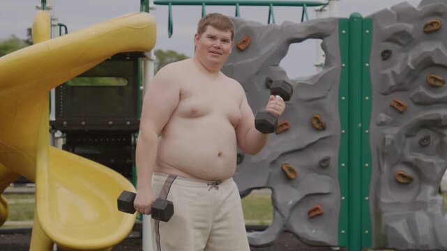 Overweight young man lifts dumbbells. Overweight fitness concept. Outdoor overweight workout uses weights. Serious overweight guy trains outside. Obesity concept with fat belly.