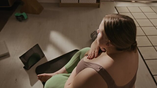Plus-size Caucasian young adult woman sitting on fitness mat at home, looking at tablet screen during workout session. Footage is ideal for body-positive wellness and online training ads