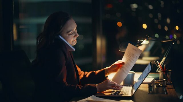 Night manager speaking cellphone sitting with laptop. Overworked businesswoman