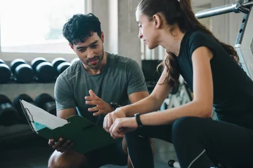 Trainer client gym clipboard consultation fitness exercise coaching session woman man discuss workout plan with focused expression near weight rack in modern health club © chokniti