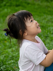 Close-up profile portrait of a happy young girl looking up with a bright, curious smile, against a soft-focus green grass background. © alenthien
