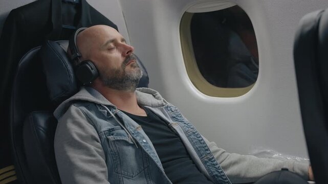 Middle-aged bald man wearing wireless headphones relaxes and sleeps in airplane cabin seat. Passenger in denim jacket rests comfortably by window during long flight with eyes closed.