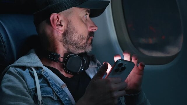Middle-aged bearded man wearing black baseball cap and denim jacket with headphones around neck uses smartphone while seated in dimly lit airplane cabin.