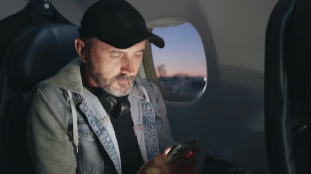 Airplane cabin scene with middle-aged man preparing for takeoff. Passenger wearing black cap and headphones around neck browses phone near window seat during dusk with sunset light visible outside.