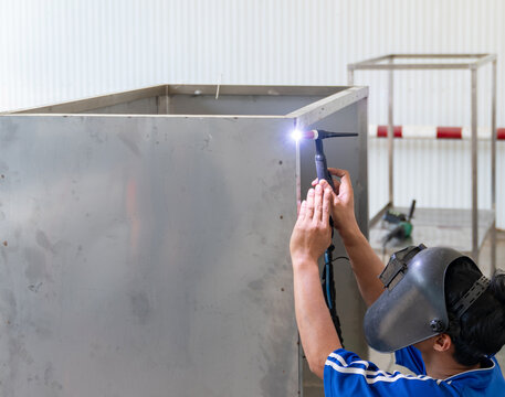 Professional welder using a TIG welding torch on a stainless steel metal box structure inside an industrial workshop.