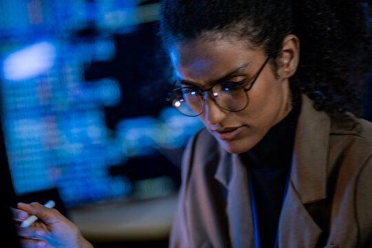 A female analyst gazes intently at data off screen, framed against glowing monitors in a dark technology room, in a professional, cutting edge technology setting.