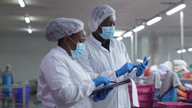 Factory quality control inspectors in protective gear checking digital data in food plant, Industrial food safety supervisors managing manufacturing process documentation