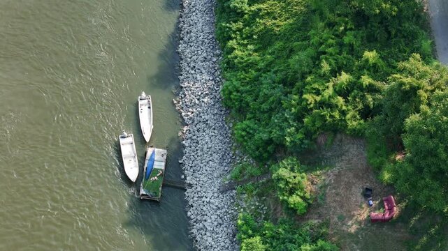 Small boats moored along the Po River beside a riprap embankment and riverside seating area near Castelvetro Piacentino, Piacenza, Emilia-Romagna, Italy, bordered by riparian woodland, static aerial