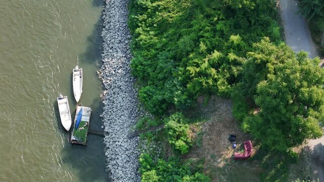 Small boats moored along the Po River beside a riprap embankment at the Maginot riverside area near Castelvetro Piacentino, Piacenza, Emilia-Romagna, Italy, with woodland and outdoor seating, static