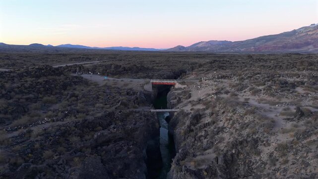 Volcanic canyon cuts through La Payunia Reserve in Mendoza, Argentina, revealing basalt formations and a bridge crossing the narrow gorge at sunrise, drone parallax pullback ascent shot.