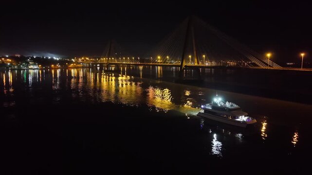Luxury catamaran yacht traversing next to the illuminated San Roque Gonzalez international bridge. Night aerial orbit at Posadas, Misiones, Argentina.