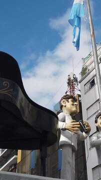 Two giant musician figures hold microphones beneath a waving Argentine flag against a Buenos Aires skyline, capturing the festive street culture and tango identity of the city, vertical pan right
