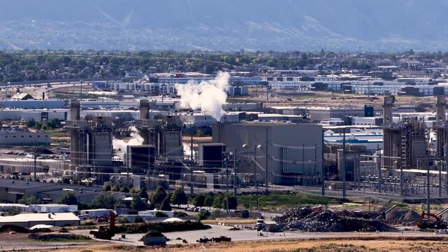 Industrial pipe coating factory featuring coating lines, curing units, cooling steam emissions from smokestacks, warehouses and utility infrastructure in Utah Valley, slow telephoto forward shot.