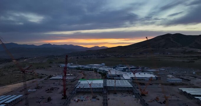 Descending aerial footage reveals a massive construction site at Silicon Slopes, Lehi, Utah, with towering cranes and Traverse Mountain silhouetted against a vibrant sunset sky.