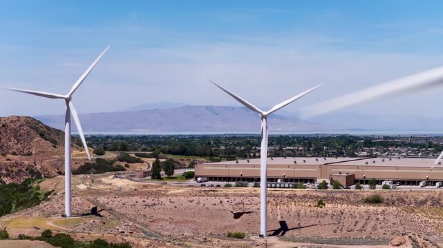 Wind turbines with three-blade rotors, nacelles and tubular steel towers above Utah Valley near Spanish Fork, Utah, USA, beneath the Wasatch Range, drone forward flyover