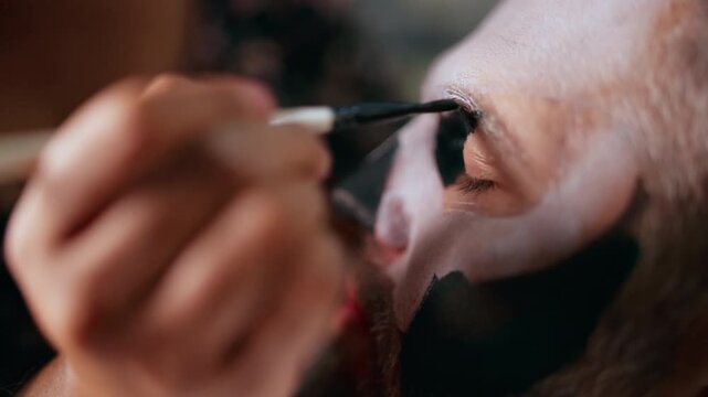 Close-up of makeup artist carefully applying dark black paint around the eye area with a fine brush, creating dramatic Day of Dead or Halloween costume makeup on bearded man with pale white face.