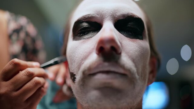 Close-up of professional makeup artist applying traditional Day of the Dead or Halloween face paint to man. White base covers face while dark black pigment surrounds closed eyes creating skull effect.