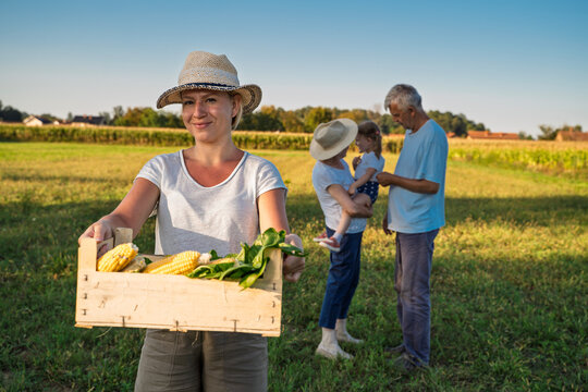 Young woman with her whole family standing on agricultural field with freshly picked organic corn in wooden crate 