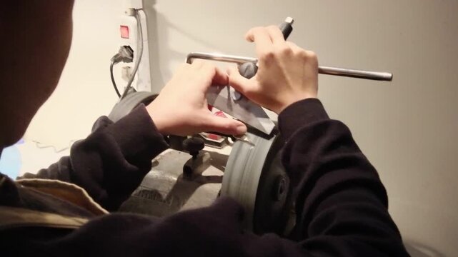 Worker sharpens a metal cutting tool on a bench grinder in an industrial workshop, producing a precision cutting edge through abrasive grinding and metalworking craftsmanship, close-up static shot.