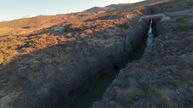 Volcanic canyon cuts through La Payunia Reserve near Malarg&uuml;e, Mendoza, Argentina, as an FPV drone races above a narrow watercourse between basalt walls and beneath a bridge, FPV ultra fast flyover