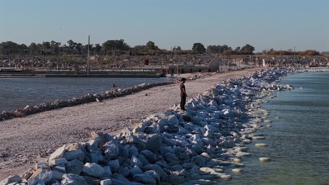 Lone tourist standing on a rocky shore next to a desolate road amidst the historical flood ruins of Villa Epecuen. Low aerial orbit during sunny and windy afternoon.