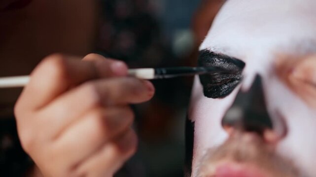 Makeup artist carefully applies black paint around the eye area creating a traditional skull design for Day of the Dead or Halloween celebration. Hand holds brush with precision.