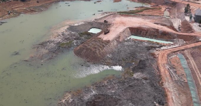 Aerial view: a construction site with excavated terrain and turquoise pools, where a truck reverses to unload earth while other machinery operates nearby.